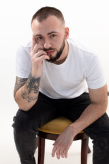 Young bearded man with short dark hair in white T-shirt, black jeans, sitting on wooden chair, leaning head on hand.