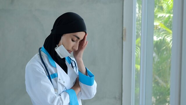 A Muslim woman doctor stands by a window in the office. Removes the medical mask from the face. He looks out the window. Touches his forehead. The Muslim woman doctor has a tired look.