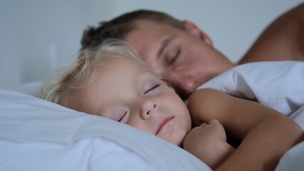 A young father, along with his daughter of two years, sleep soundly next to each other in bed on white bedding. They have deep dreams. They sleep in a bright cozy room. Father guards baby's sleep