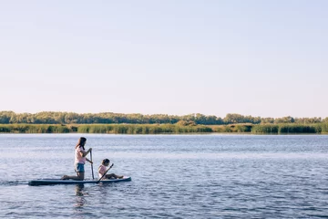 Foto op Canvas Mother and small daughter paddle boarding together on one sup on lake rowing with oars with reeds and blue sky in background. Active lifestyle. Teaching children to love sports from childhood. © Юля Бурмистрова