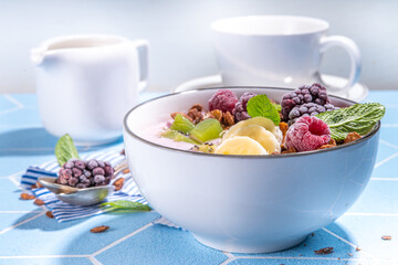 Summer breakfast healthy diet bowl with yogurt, granola, fresh fruit and berries, One portion with overnight fruity granola oats on sun-lighted tiled blue background copy space