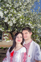 Young loving couple man and woman in white embroidered shirts near a flowering tree in a spring garden.