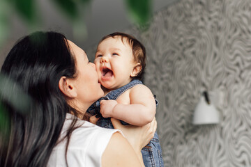 Portrait of young mother with little daughter in bedroom closeup, free copy space, blurred background and green plants. Hold in hand screaming baby. Concept of maternal affection and childcare.