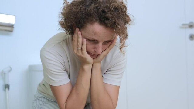In the toilet room, where the toilet and other sanitary devices are located, a woman sits on a floor toilet in clothes with a tired look and thinks about something. Women's problems and diarrhea.