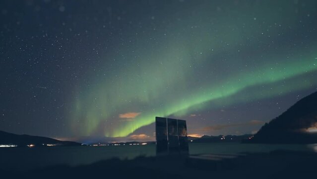 Aurora borealis above the Battle for Narvik memorial on the shore of the fjord. Street and car lights flash in the background. Timelapse.