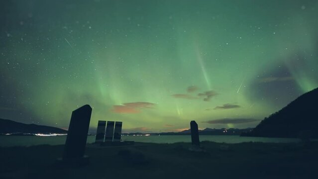 Northern lights above the Battle for Narvik memorial on the shore of the fjord. Street and car lights flash in the background. Timelapse.