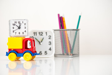 on a white table in front of a blurry clock and a glass with pencils there is a toy car with a small clock on top