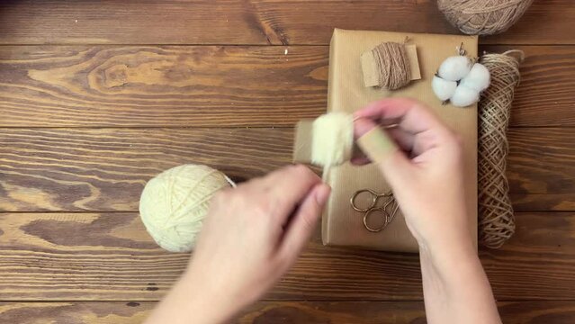Women's hands wind white yarn on piece of cardboard. Box in craft wrapper, twine, cotton and scissors on wooden table. Handmade crafting decorations of gift for Christmas or Birthday. Festive mood.