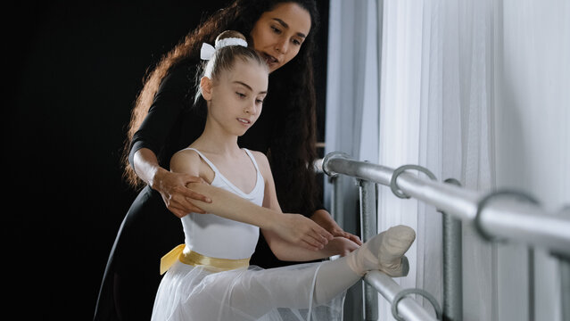 Female choreographer teacher mentor trainer of classical dance teaches posture help with stretching to young student learn dancing teen girl ballerina near ballet barre. Teaching gymnastics class
