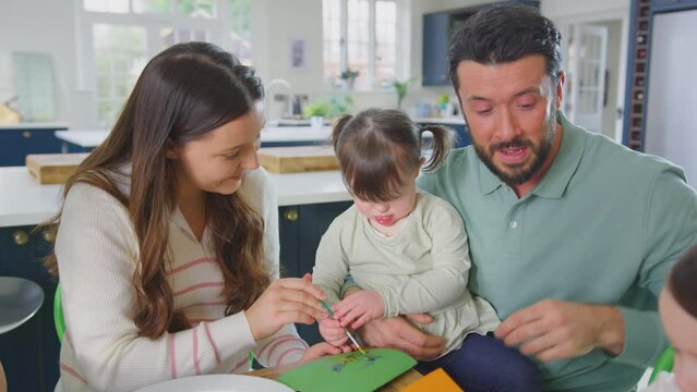 Family with Down Syndrome daughter sitting around table at home having fun using paint and pencils to make homemade greeting card together - shot in slow motion