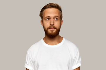 Head and shoulders image of brutal handsome pensive Caucasian ginger-head male dressed in white t-shirt with blank copy space for your advertising text, standing over grey wall, looking aside