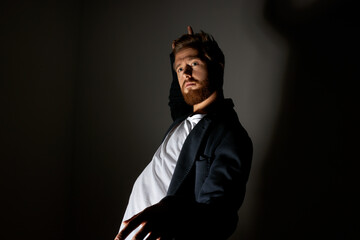 Male fashion. Portrait of charismatic photogenic ginger-headed man with beard and moustache posing in black trendy coat at studio against dark background casting shadow on wall. Light and shadow