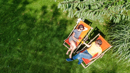 Young couple relax in summer garden in sunbed deckchairs on grass, woman and men have drinks on...