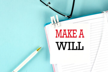 MAKE A WILL text on a sticky on notebook with pen and glasses , blue background