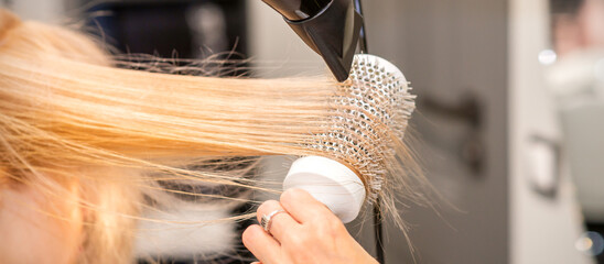 Fototapeta premium Drying straight blond hair with black hairdryer and white round brush in hairdresser salon, close up