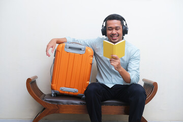 Adult Asian man sitting with travelling suitcase beside while listening music and read a book - obrazy, fototapety, plakaty