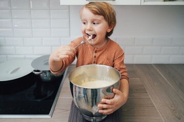 Cute little blond preschool kid boy baking cake in domestic kitchen, indoors. Funny child  tasting...