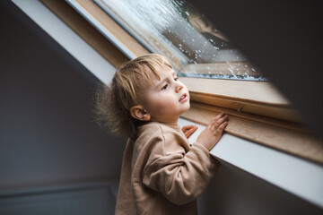 Little preschool boy staying home in bad weather and looking  with interest through window on...