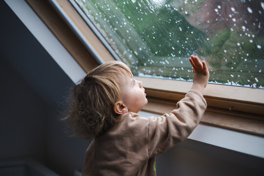 Little preschool boy staying home in bad weather and looking  with interest through window on raindrops and hail, idoors.