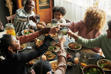 African big family toasting with glasses with lemonade while having holiday dinner together at table at home - Powered by Adobe