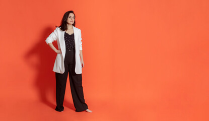 Young brunette woman in black trouser suit and white jacket stands on orange background. Looks into...