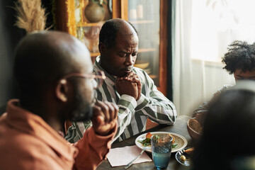 African mature man sitting at table with his eyes closed and praying with his family during dinner