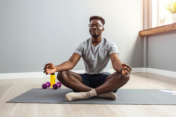 Man in sporty outfit doing yoga and meditating on an exercise mat. Sporty mindful man with tattoo meditating alone at home, peaceful calm hipster fit guy practicing yoga in lotus pose - obrazy, fototapety, plakaty