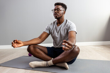 Man in sporty outfit doing yoga and meditating on an exercise mat. Sporty mindful man with tattoo meditating alone at home, peaceful calm hipster fit guy practicing yoga in lotus pose