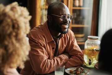 African happy man in eyeglasses sitting at dining table talking and laughing with his family