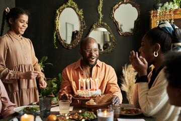 Happy African man sitting at table with birthday cake and celebrating birthday with his family at...