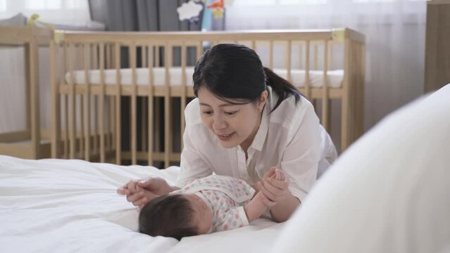 joyful young mother leaning by the bedside is smiling and interacting with her newborn child lying on the bed and holding its cute little hands.
