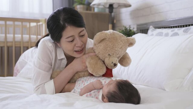cheerful first time mother lying prone by bedside is talking to her young child with a teddy bear in hands at the bright home interior.