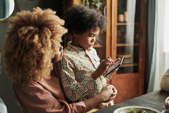 African little son using mobile phone while sitting on knees of his mother while they having dinner at table