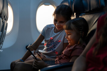 Adorable little girls travel by plane. Cute Kids sit at the window of an airplane and use a digital...