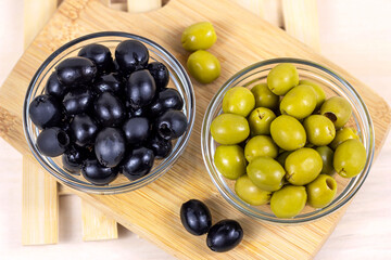 Top view of many green and black marinated whole olives in the glass bowls on light wooden board background in the kitchen.