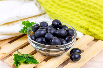 Many black marinated whole olives in the glass bowl on light wooden board background in the kitchen.