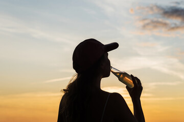 Fototapeta premium Silhouette of girl in baseball cap drinking soda water from glass bottle and looking at sunset sky. Rear view of female