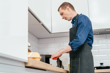 Portrait of middle-aged man wearing blue long-sleeve shirt and black apron chopping red bell pepper with knife on wooden cutting board on kitchen table. Cooking, healthy food.