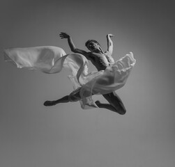 Liberty. Black and white portrait of graceful muscled male ballet dancer dancing with fabric, cloth isolated on grey studio background. Grace, art, beauty concept.