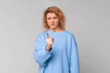 Serious middle age woman with curly blonde hair is warning, shakes her finger threateningly, standing in trendy blue sweatshirt over light grey background