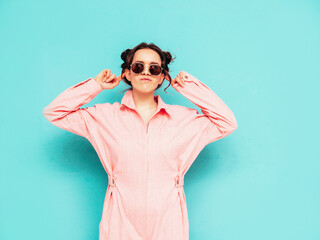 Young beautiful smiling female in trendy summer pink overalls. Sexy carefree woman posing near blue wall in studio. Positive model having fun and going crazy. Cheerful and happy. Dancing in sunglasses