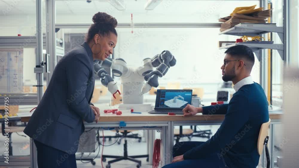 Poster black senior engineer talks to young male computer scientist while analysing robotic arm at the rese