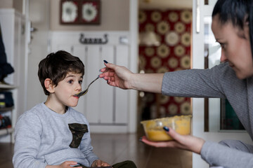 Mother feeds puree to her small disabled son sitting on the floor while the child plays with the mobile phone