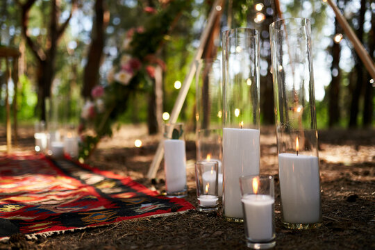 Candles burning in glass vases, flasks on carpet near bohemian tipi arch decorated with roses and flowers wrapped in fairy lights on outdoor wedding ceremony venue in pine forest. Boho rustic decor.