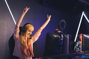 Fototapeta na wymiar Victory. Excited satisfied long-haired female gamer influencer wins her next online match in a tournament. Happiness shown by throwing hands in the air. Internet cafe. High quality photo