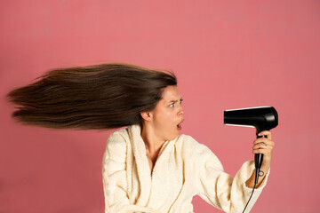 Funny woman drying her long hair with electric fan on a pink background