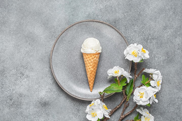 Ice cream in a waffle cone on a plate with a decorative cherry blossom branch. Gray concrete grunge...