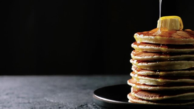 Honey or maple syrup is poured over a stack of pancakes with butter moving on dark background. High quality 4k footage