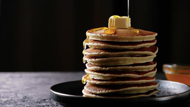 Honey or maple syrup is poured over a stack of pancakes with butter on dark background. Close-up high quality 4k footage