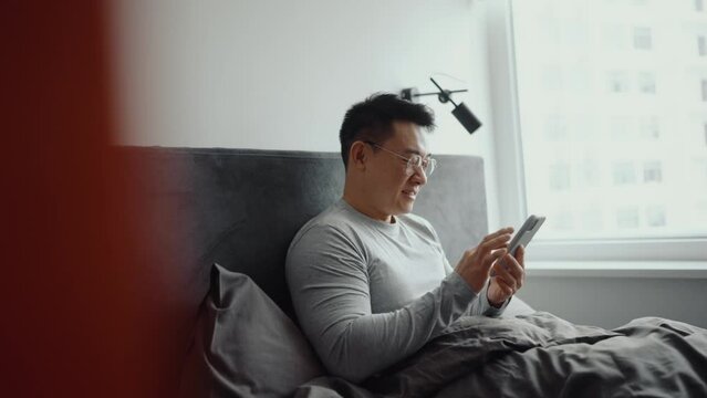 Smiling Asian man wearing eyeglasses typing by mobile on the bed at home